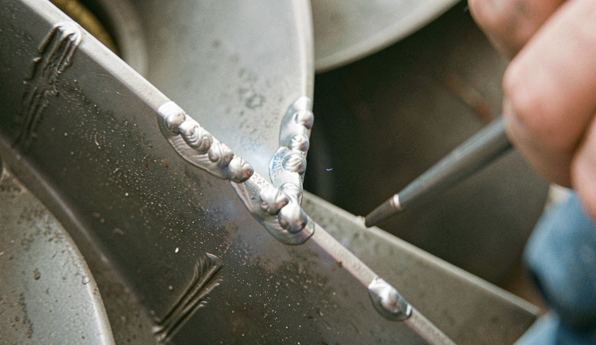 Close view of a fan blade leading edge with hardfacing weld beads applied, wear grooves visible in the parent plate, a welder's glove resting on the rotor